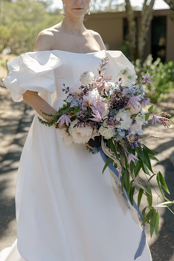 Bridal portrait of a bride in an off the shoulder wedding dress holding a pastel bouquet with blue ribbon on a sunlit walkway lined with trees