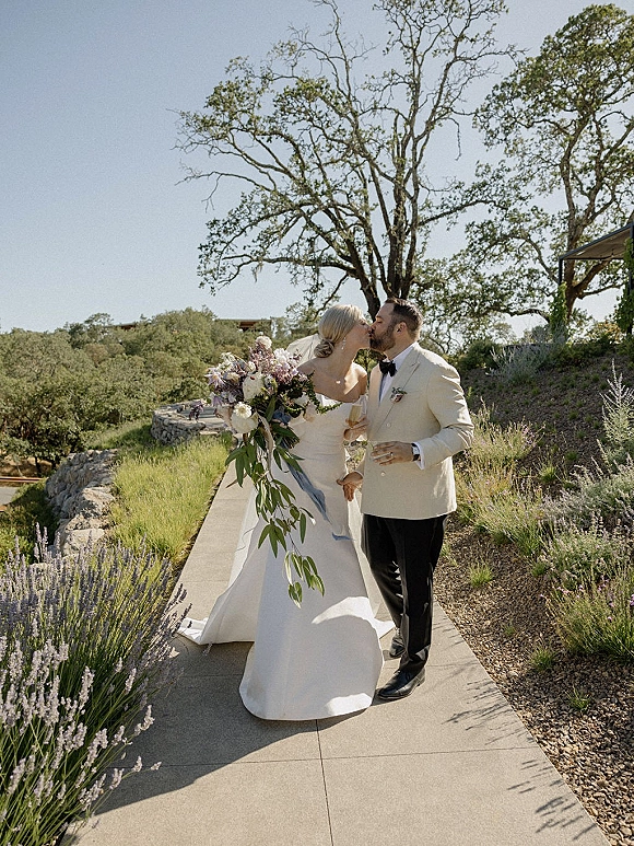 Wedding kiss portrait of bride and groom kissing on an outdoor walkway, bride holding bouquet with veil as lavender and stone wall line the path
