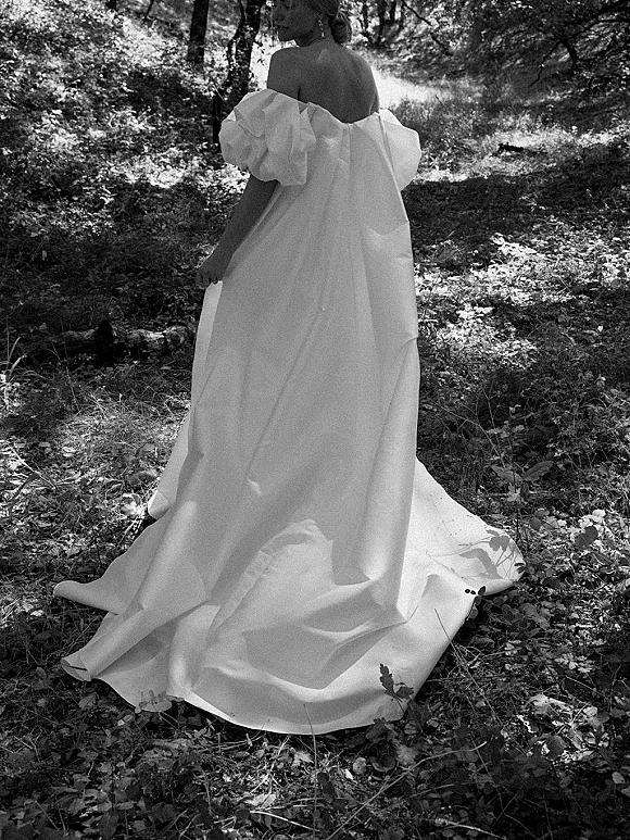 Bridal portrait of a bride from behind walking a woodland path in dappled sunlight, wearing an off the shoulder wedding dress with train