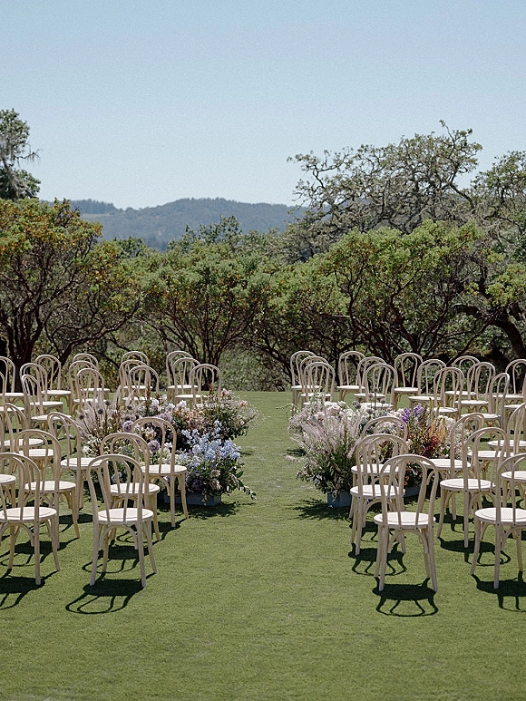 Ceremony seating with outdoor wedding ceremony chairs in neat rows, flanked by meadow grass aisle and pampas floral clusters on a lawn with hills