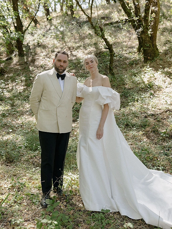 Couple portrait of bride in an off shoulder wedding dress with train and groom in a double breasted tuxedo in dappled forest light