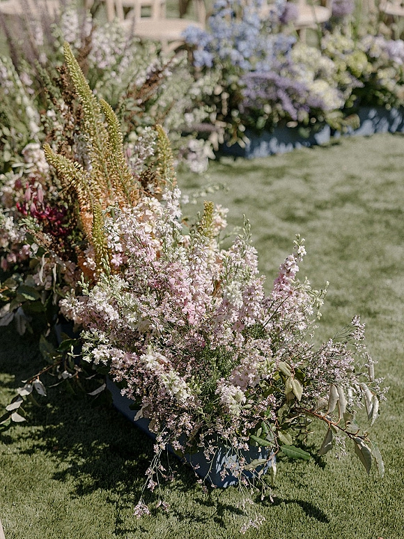 Wedding floral arrangement of pastel wildflowers and greenery in blue planters lining a garden ceremony aisle on a grass lawn with white chairs