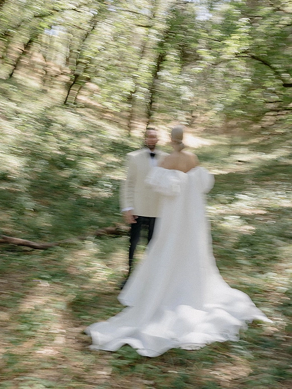 Couple portrait of bride in strapless gown with long train and groom in white tuxedo jacket on a sunlit forest path