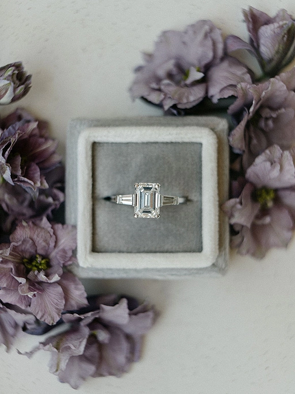Engagement ring, emerald cut engagement ring resting in a gray velvet ring box with lilac flowers on a light stone surface