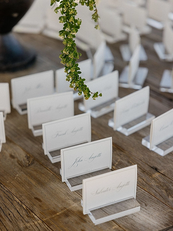 Wedding place cards with script calligraphy in clear holders, styled with greenery accents on a rustic wood table at the reception