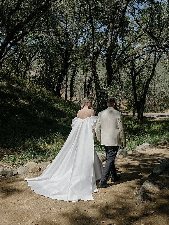 Couple portrait of bride and groom walking away holding hands, her off-the-shoulder dress train trailing on a sun-dappled woodland path