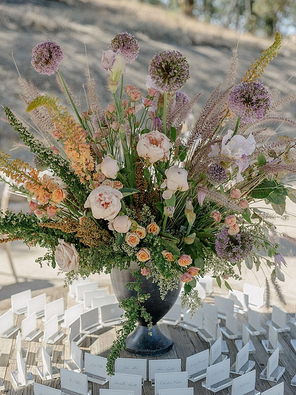 Wedding floral centerpiece in a large urn vase with roses, peonies, and allium, surrounded by place cards on a wooden table outdoors