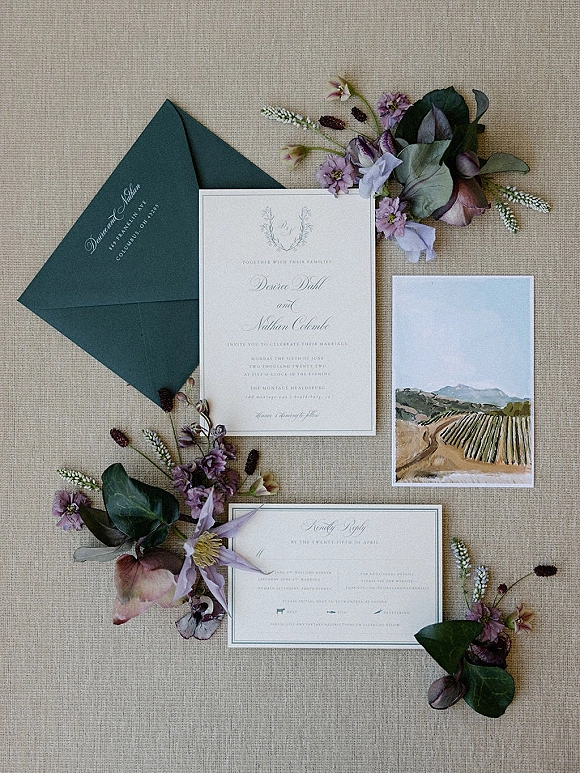 Invitation suite wedding invitation flat lay with dark green envelope, calligraphy cards, wax seal, and purple sprigs on linen backdrop