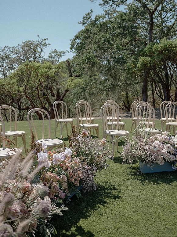 Ceremony setup with outdoor wedding ceremony seating, bentwood chairs lining a grassy aisle with pastel rose florals under blue sky