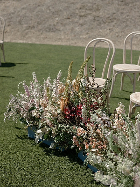 Ceremony aisle flowers create a wildflower meadow border with pastel blooms and greenery beside white bentwood chairs on a grassy lawn