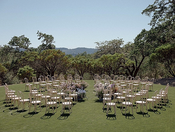 Ceremony setup with outdoor wedding ceremony chairs in a circle, wood seating and meadow aisle florals on a lawn under oak trees