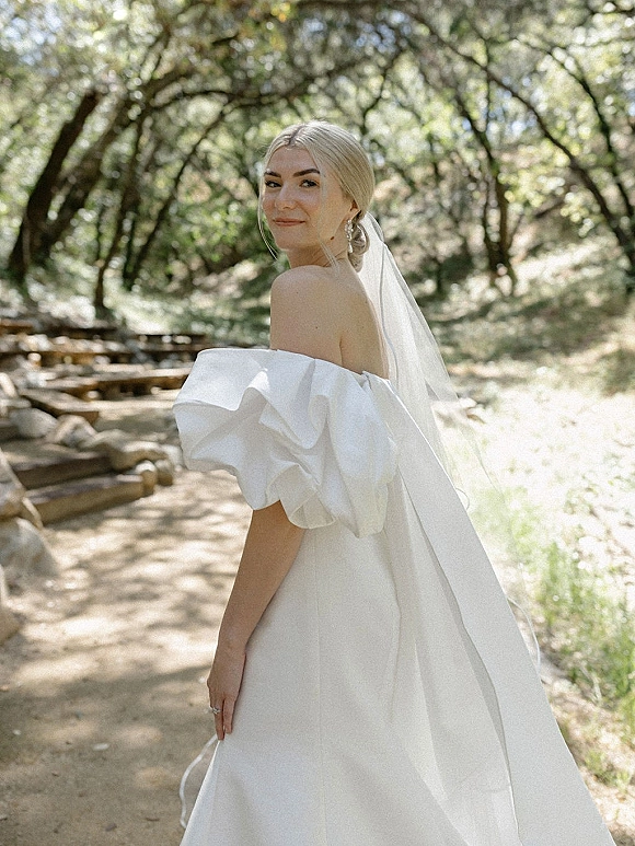 Bridal portrait of a bride in a strapless wedding dress with a long veil, looking back on a woodland path in dappled sunlight