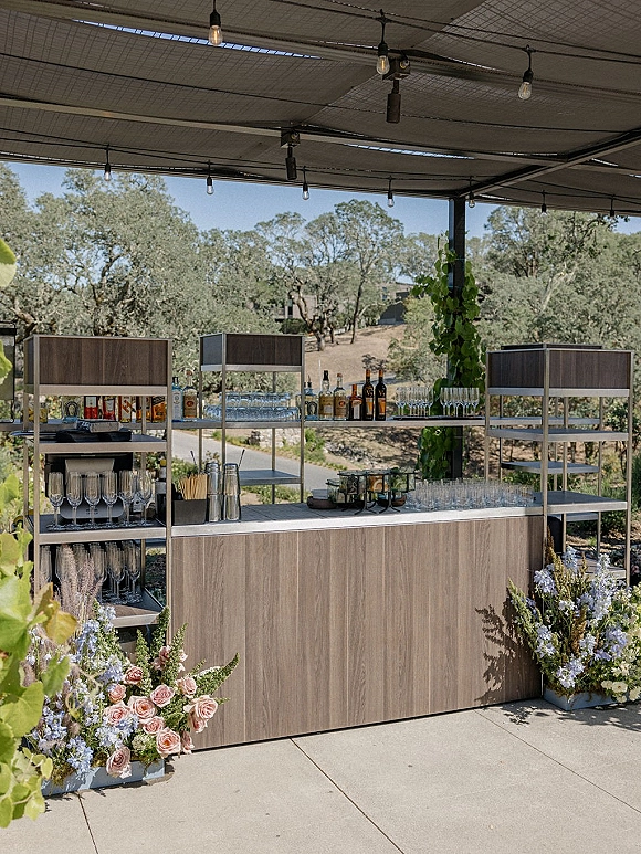 Wedding bar setup on a wood bar with open shelving, bottles and glassware, accented by greenery garland under patio string lights outdoors