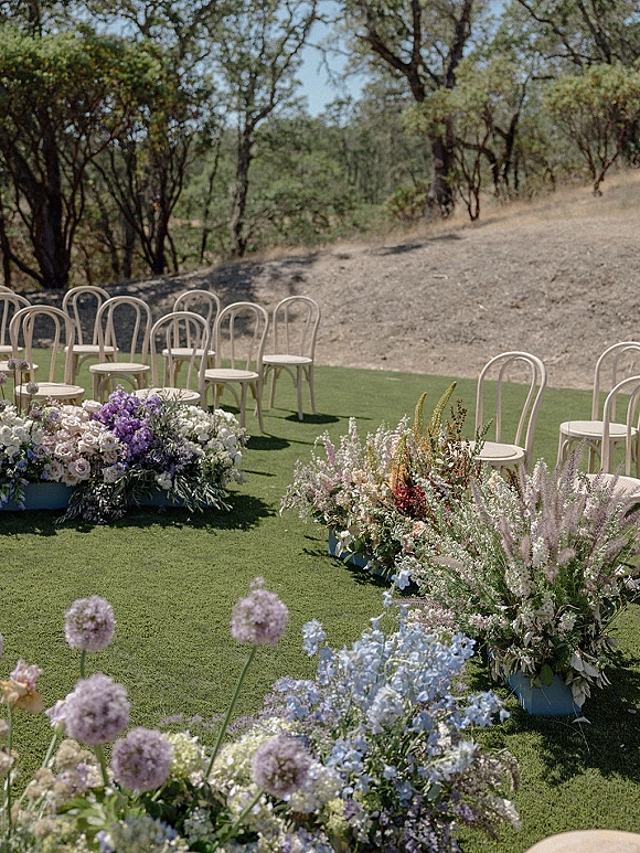Ceremony aisle decor with ground floral aisle clusters of pastel meadow flowers and greenery beside bentwood chairs on a lawn hillside