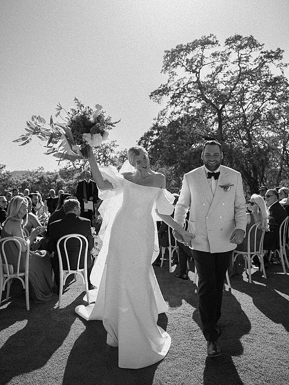Wedding recessional as bride and groom walk the aisle holding hands, bouquet raised, veil flowing, guests cheering on a sunny lawn
