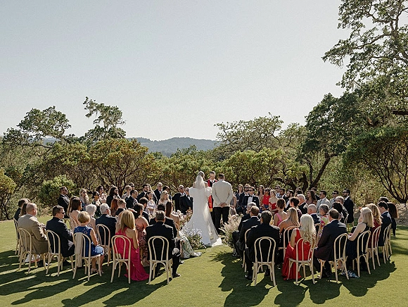 Outdoor wedding ceremony with bride in white gown and long veil beside groom and officiant, guests seated in a circle on a grassy lawn with trees and hills