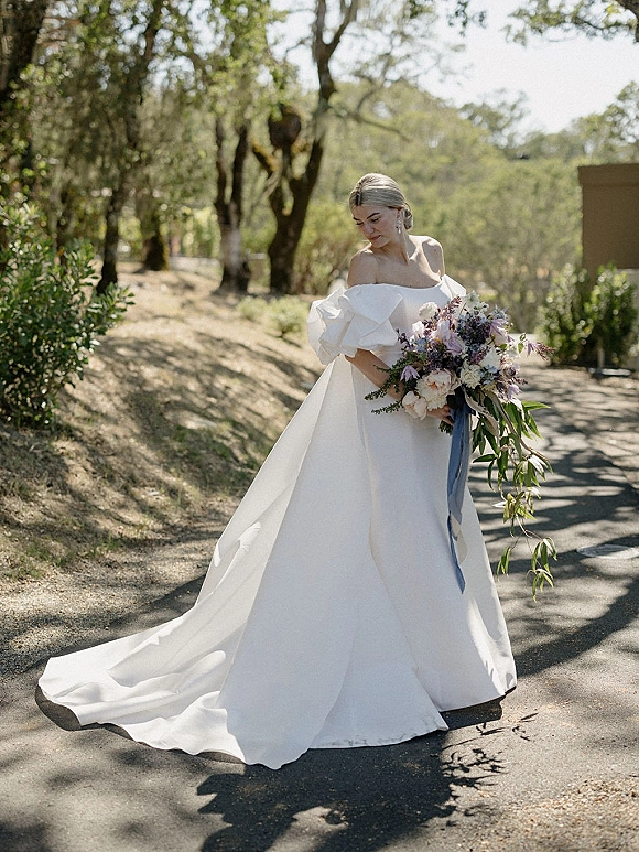 Bridal portrait of a bride in an off the shoulder wedding dress with long train, holding a bouquet with blue ribbon on a sunlit road by trees