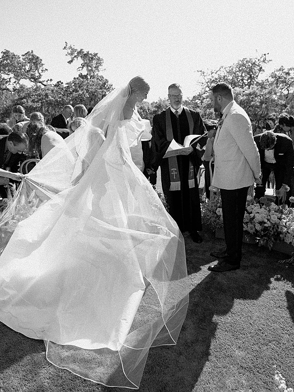 Wedding ceremony with couple exchanging vows as bride’s veil trails behind her, bouquet in hand, on an outdoor lawn with guests and flowers