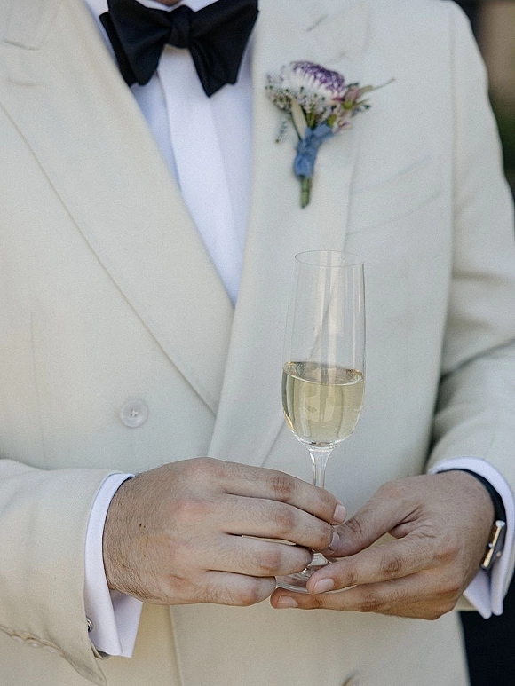 Groom portrait in a white tuxedo jacket, holding a champagne flute with boutonniere, cufflinks, and wristwatch against blurred greenery