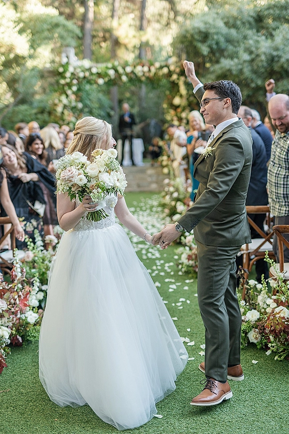 Ceremony recessional as newlyweds walk the aisle holding hands, groom fist pumping, rose petals on green runner in garden setting