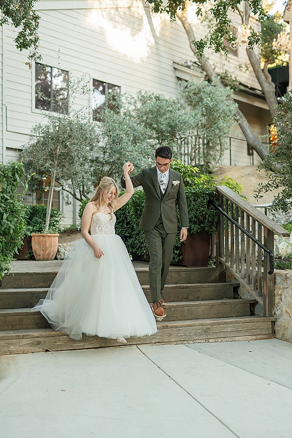Couple portrait of bride and groom walking hand in hand down outdoor stairs, groom in green suit and bride in strapless lace dress train