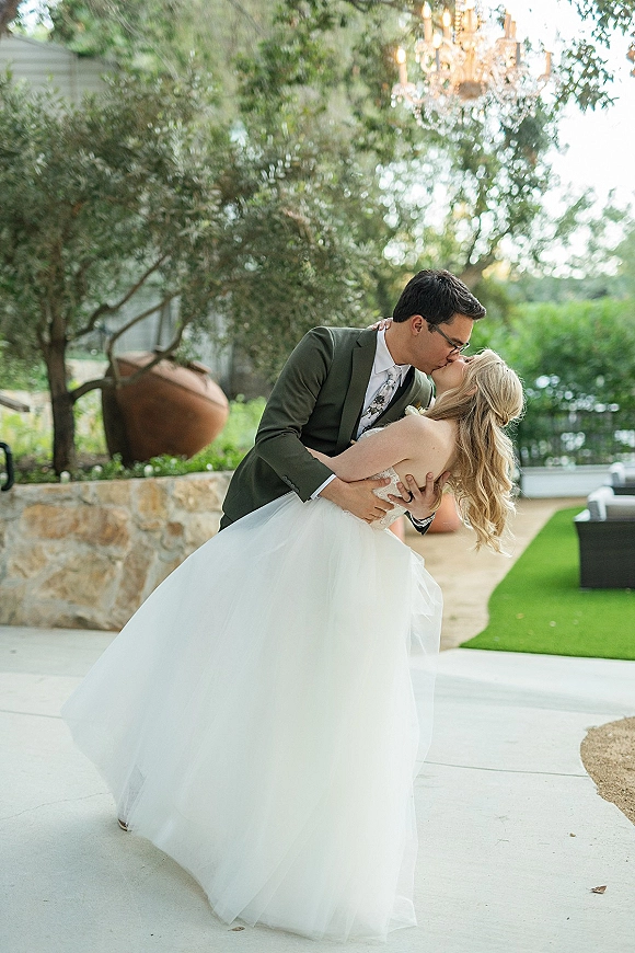 Wedding kiss portrait of groom dipping bride in a dip kiss pose, her tulle ball gown flowing on a garden patio with chandelier