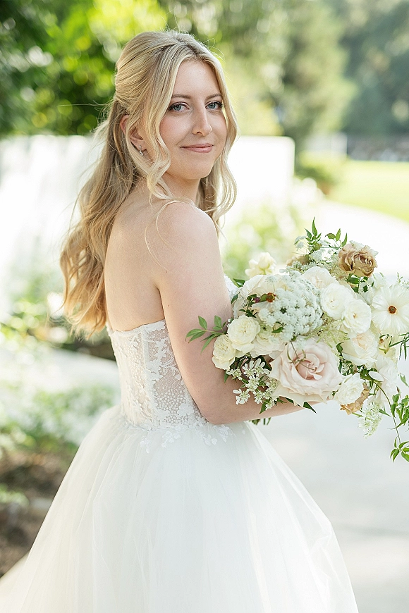 Bridal portrait of a bride holding bouquet in a strapless lace dress, looking over her shoulder in a lush garden setting