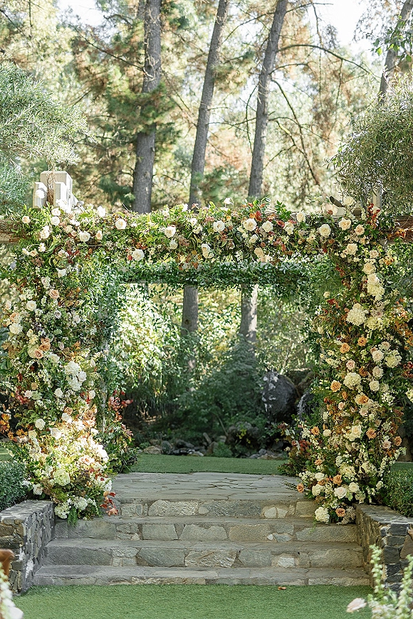 Wedding ceremony altar framed by a floral ceremony arch with rose and greenery garland on stone steps, set against garden trees and lawn