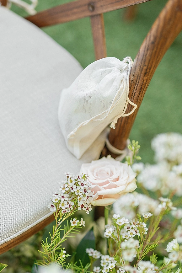 Wedding chair detail with ceremony chair decor, a white drawstring favor bag tied to a wooden chair with blush rose and waxflower on a green lawn