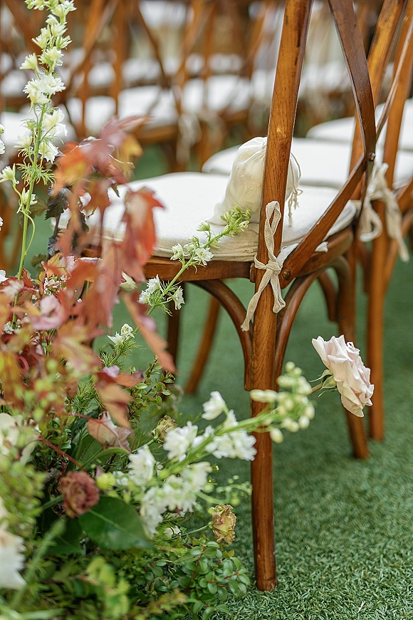 Ceremony aisle decor with outdoor ceremony chairs, cream cushions and fabric ties, plus low florals and greenery on a green lawn
