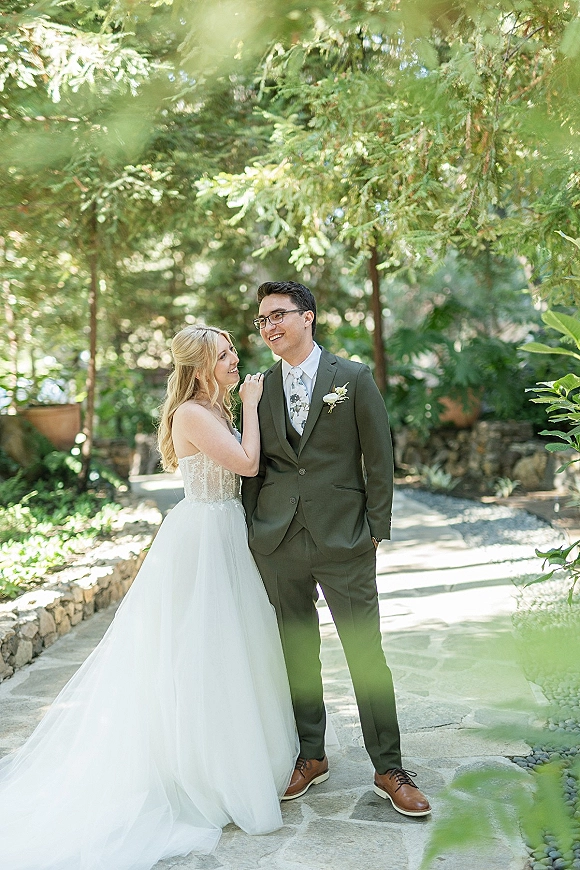 Couple portrait of bride and groom smiling as she leans on him, groom in green suit with eyeglasses on a stone garden path