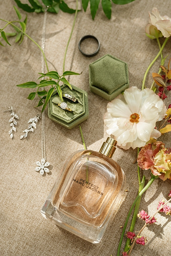 Wedding rings in a green velvet ring box with engagement ring flat lay on linen, surrounded by bridal jewelry, perfume, and greenery