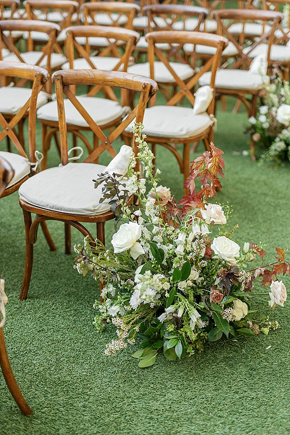 Ceremony aisle decor with aisle floral arrangements of white roses, greenery and autumn leaves beside cross back chairs on a grass lawn