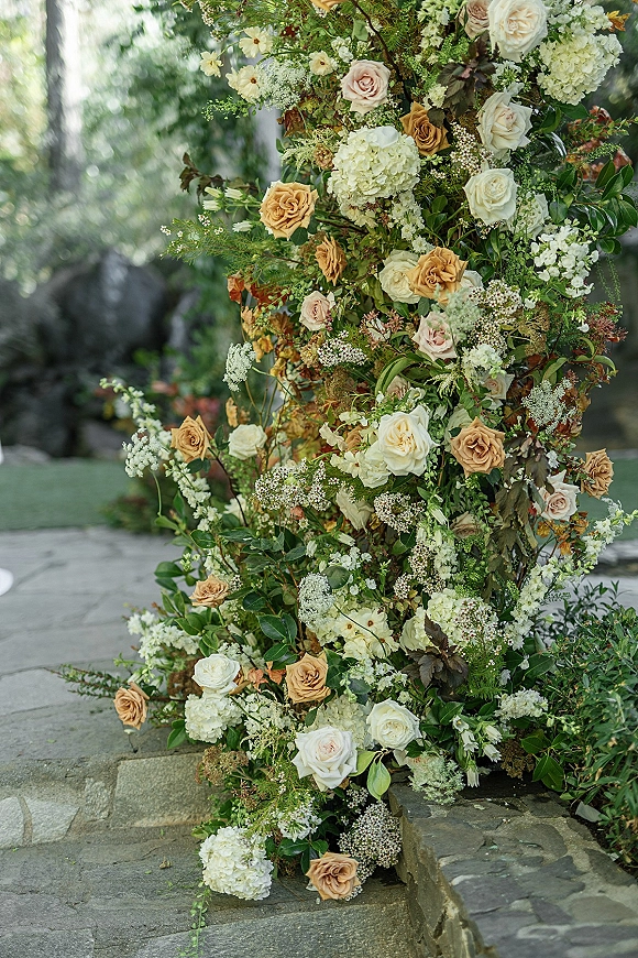 Wedding floral installation with a ceremony floral pillar of roses and hydrangeas with greenery on a stone patio amid garden foliage
