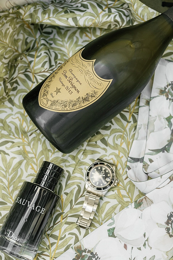 Wedding champagne bottle in a wedding detail flat lay with cologne bottle and wristwatch on a green botanical print surface