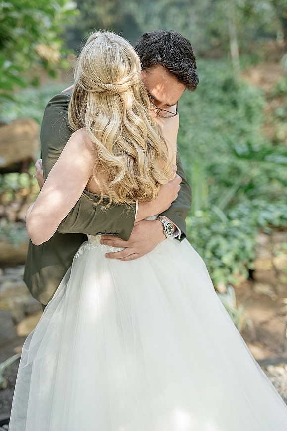 Wedding hug as newlyweds embracing, bride in strapless lace bodice and tulle skirt holding groom in glasses on a garden stone path