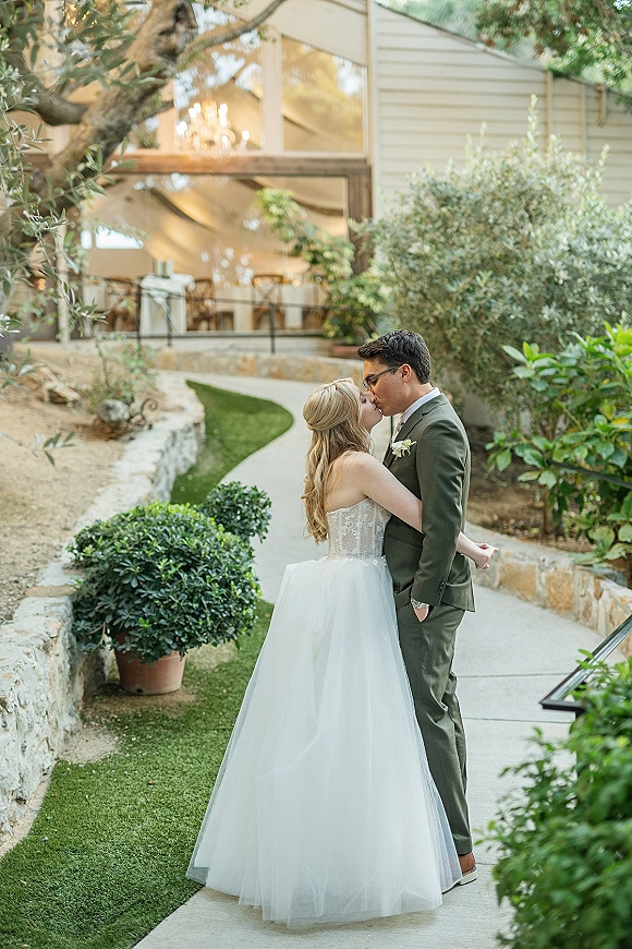 Wedding kiss as bride in a strapless lace dress and groom in an olive green suit embrace on a garden walkway near a stone wall