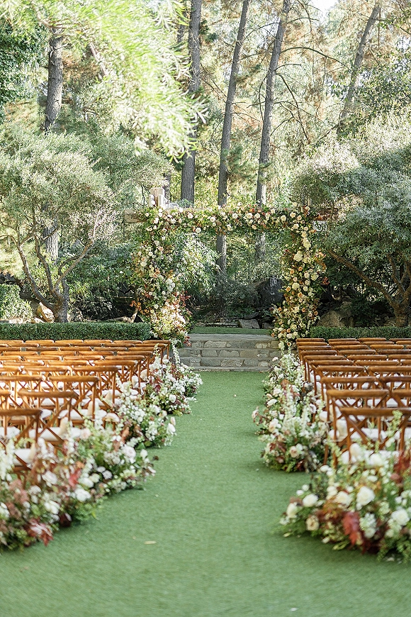 Ceremony setup for an outdoor wedding ceremony with a floral arch, aisle flowers, and wooden chairs on stone steps amid garden trees