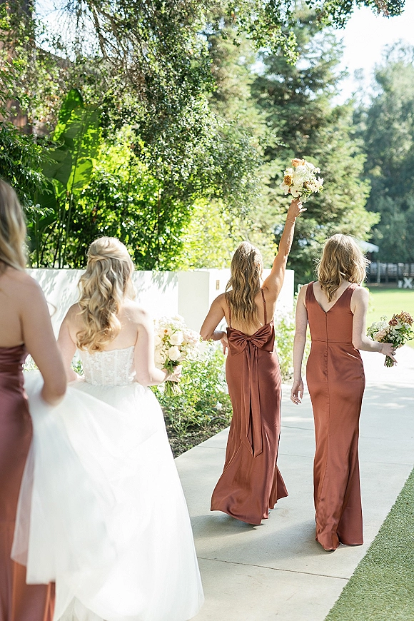 Bridesmaid moment as bridesmaids walking away with bouquets, one holding hers up, beside the bride on a garden walkway