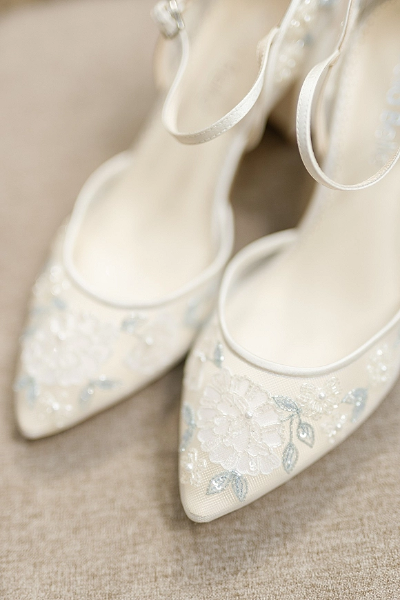 Wedding shoes with bridal heels styling, ivory pointed-toe ankle-strap pumps with floral lace applique and beading on neutral fabric surface