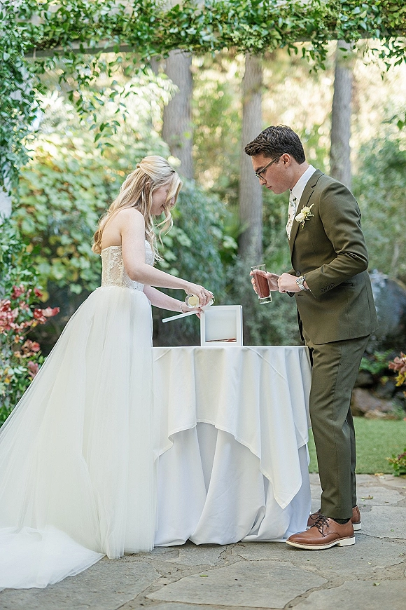 Unity ceremony as bride in lace bodice gown and groom in green suit pour wine into a unity box on a white linen table outdoors