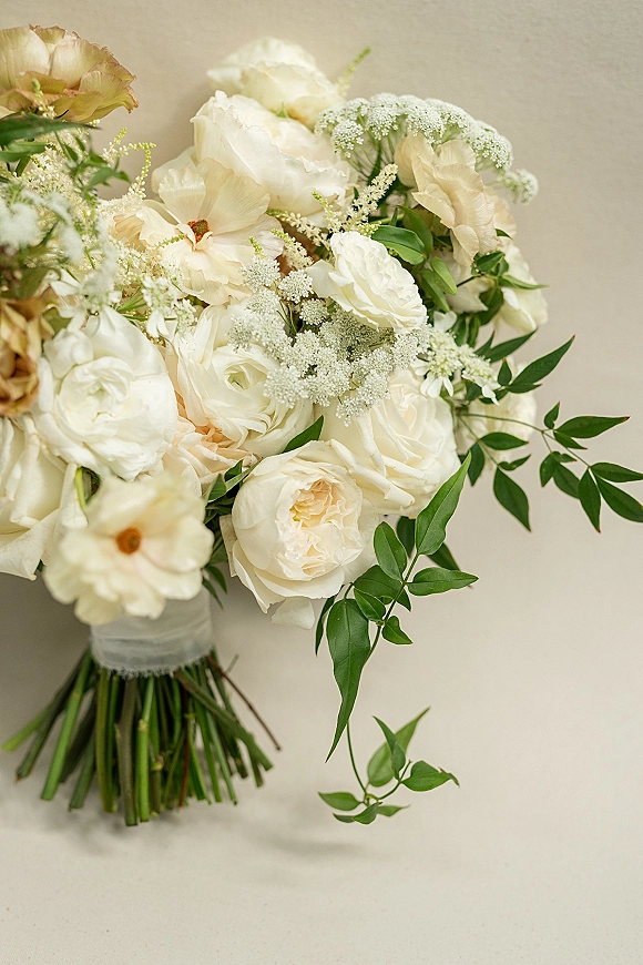 Bridal bouquet of white rose bouquet blooms with peonies, ranunculus, baby’s breath and greenery, wrapped with ribbon on a neutral wall backdrop