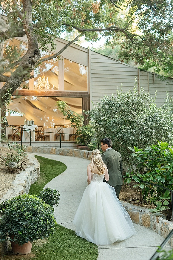 Couple portrait of bride and groom walking away along a garden path, her veil trailing, heading toward a glass-walled reception space with chandelier