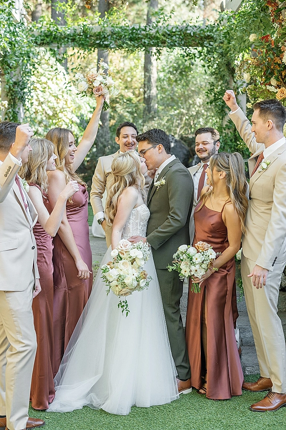 Wedding party portrait of bride and groom kissing as bridesmaids and groomsmen cheer, bride holding bouquet under a greenery arch outdoors