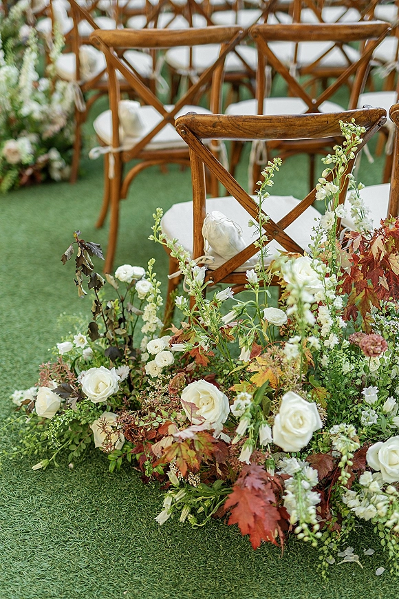 Ceremony aisle florals with grounded aisle flowers of white roses, greenery, and autumn leaves beside cross back chairs on a grass lawn