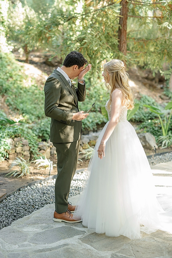 First look moment as bride approaches groom in a garden, his emotional reaction with suit boutonniere beside a stone path