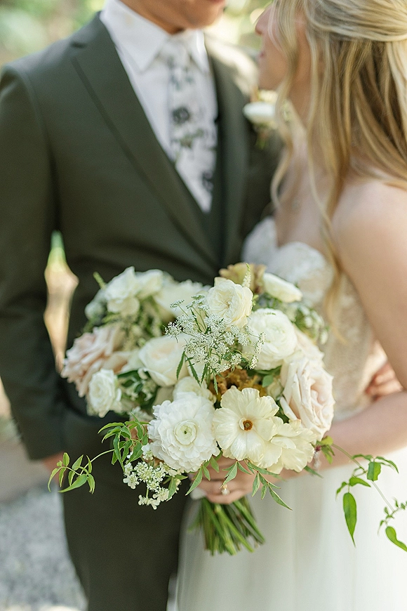 Bridal bouquet of white rose and blush blooms with ranunculus and greenery held by bride and groom in soft outdoor light