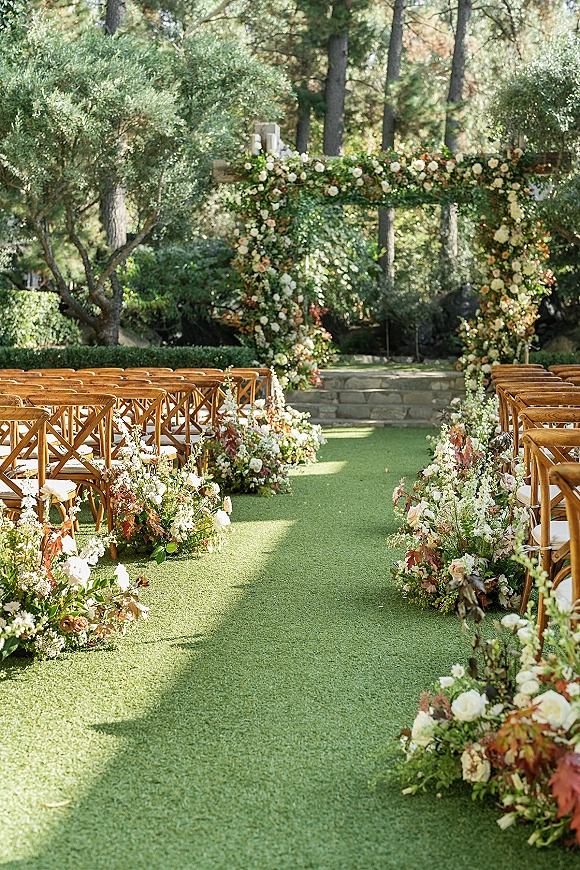 Ceremony setup for outdoor wedding ceremony with floral arch and rose-lined aisle, crossback chairs on a garden lawn by stone steps