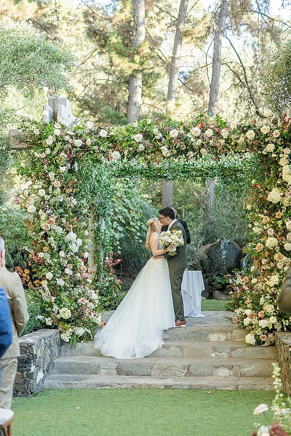 Ceremony kiss as the bride and groom embrace under a floral arch, with a bridal bouquet and guests on stone steps in a garden setting