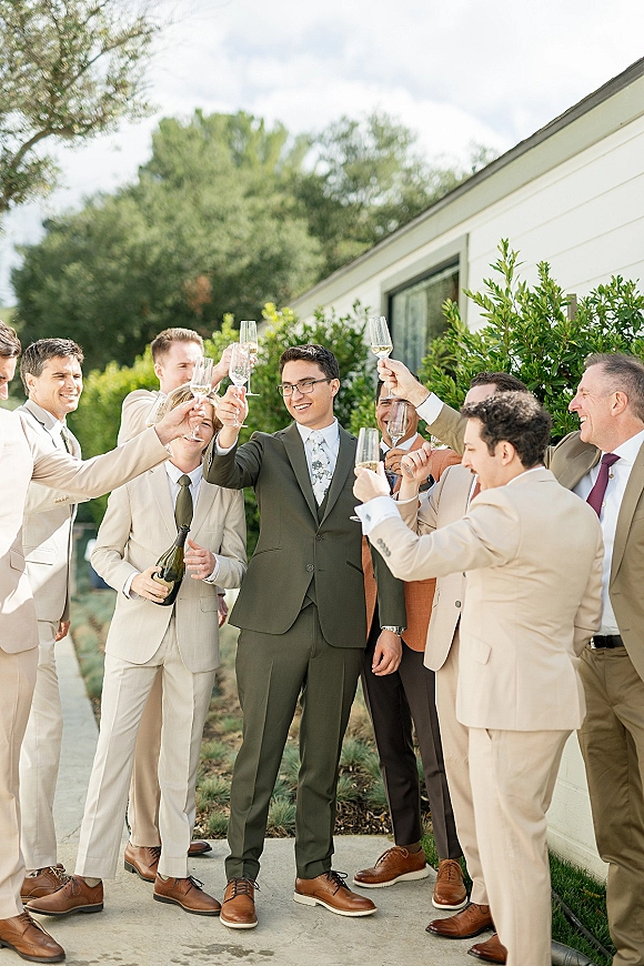 Groomsmen toast with champagne flutes as the groom and friends in tan suits cheer on a sidewalk by shrubs and trees outdoors